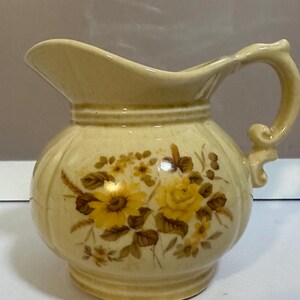 Può includere: Brocca in ceramica color crema con corpo arrotondato e manico curvo. La brocca è decorata con disegni floreali gialli e marroni. Il beccuccio è delicatamente curvo e il manico ha un ricciolo decorativo alla base.