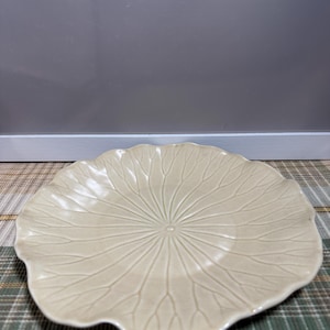 May include: A round, cream-colored ceramic plate with a scalloped edge and a raised, radiating vein pattern that resembles the structure of a leaf. The surface has a glossy finish and a central point from which the symmetrical lines extend outward.