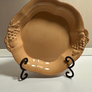 May include: A tan-colored ceramic serving plate with a wavy edge and grape cluster details. The plate is displayed on a black metal stand, suitable for serving appetizers or desserts. The plate has a glossy finish.