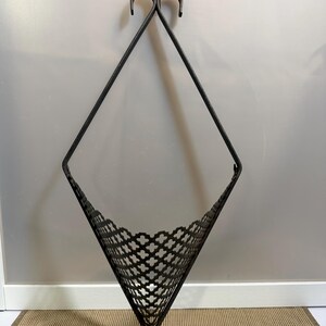 May include: A dark brown metal wall-mounted basket with a diamond-shaped frame and a perforated lattice pattern. The structure features two curved hooks at the top for hanging and two smaller curved hooks at the bottom base of the basket.