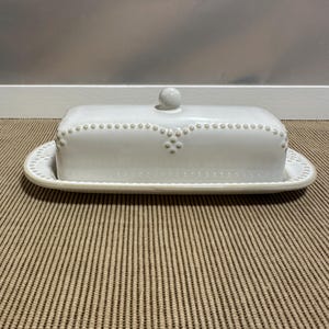 以下が含まれることがあります： 長方形のベースと蓋が付いた白い陶器製のバターケース。蓋の中央上部には小さな丸い取っ手があり、縁には盛り上がったビーズ模様、側面にはスカラップ状のデザインが施されています。