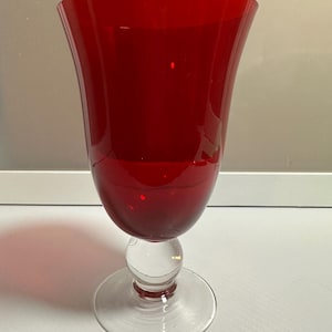 May include: A tall, ruby red glass goblet with a flared top and a clear, round base. The goblet has a bulbous midsection and a clear, circular foot. The stem is a clear, spherical shape. The glass is smooth and reflects light.