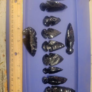 May include: A collection of black obsidian arrowheads of various sizes and shapes, arranged in a blue tray next to a wooden ruler. The arrowheads are highly polished and reflect light, showcasing their dark, glassy appearance.