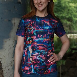 K&ouml;nnte beinhalten: Kurz&auml;rmeliges T-Shirt mit einem lebendigen, farbenfrohen Stadtlandschaftsdesign. Das Shirt zeigt eine Vielzahl von Geb&auml;uden und Strukturen in Blau-, Rosa- und Lilat&ouml;nen. Die Worte "City Market" sind auf dem Shirt zu sehen.