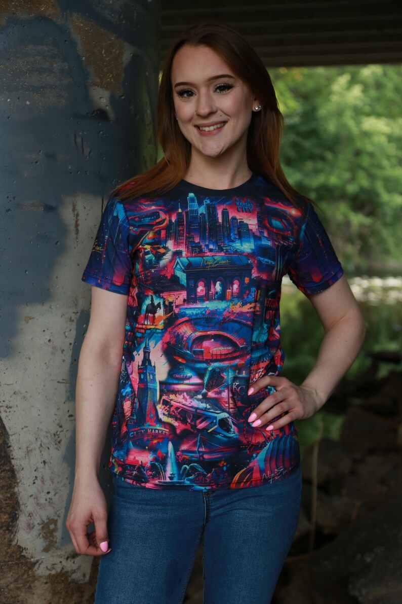 K&ouml;nnte beinhalten: Kurz&auml;rmeliges T-Shirt mit einem lebendigen, farbenfrohen Stadtlandschaftsdesign. Das Shirt zeigt eine Vielzahl von Geb&auml;uden und Strukturen in Blau-, Rosa- und Lilat&ouml;nen. Die Worte "City Market" sind auf dem Shirt zu sehen.