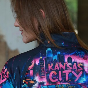 May include: A black zip-up jacket featuring a vibrant, colorful graphic of the Kansas City skyline at night. The design includes the words "Kansas City" in large, stylized letters, and the artist's signature.