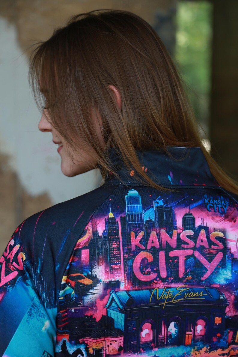 May include: A black zip-up jacket featuring a vibrant, colorful graphic of the Kansas City skyline at night. The design includes the words "Kansas City" in large, stylized letters, and the artist's signature.