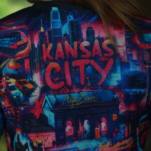 K&ouml;nnte beinhalten: Ein farbenfrohes T-Shirt mit einem lebendigen Stadtbild-Design und dem Text "Kansas City" in gro&szlig;en, roten Buchstaben. Das Kunstwerk zeigt Geb&auml;ude, Autos und einen Zug, mit der Signatur des K&uuml;nstlers "Nate Evans".