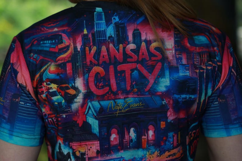 K&ouml;nnte beinhalten: Ein farbenfrohes T-Shirt mit einem lebendigen Stadtbild-Design und dem Text "Kansas City" in gro&szlig;en, roten Buchstaben. Das Kunstwerk zeigt Geb&auml;ude, Autos und einen Zug, mit der Signatur des K&uuml;nstlers "Nate Evans".