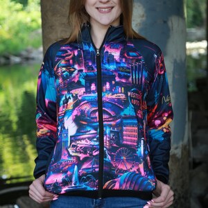 May include: A zip-up jacket with a vibrant cityscape design in shades of pink, blue, and purple. The jacket features a black zipper and a black collar. The jacket is worn over blue jeans.