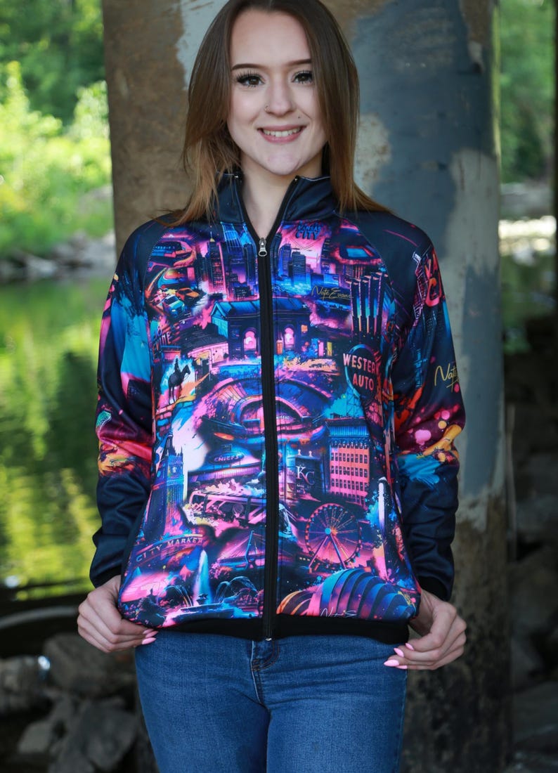 May include: A zip-up jacket with a vibrant cityscape design in shades of pink, blue, and purple. The jacket features a black zipper and a black collar. The jacket is worn over blue jeans.