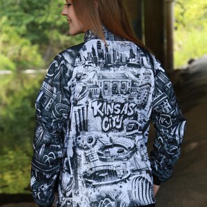 May include: A black and white zip-up jacket featuring a detailed graphic of Kansas City landmarks and the city's name. The jacket has a black collar and cuffs, and is worn with blue jeans.