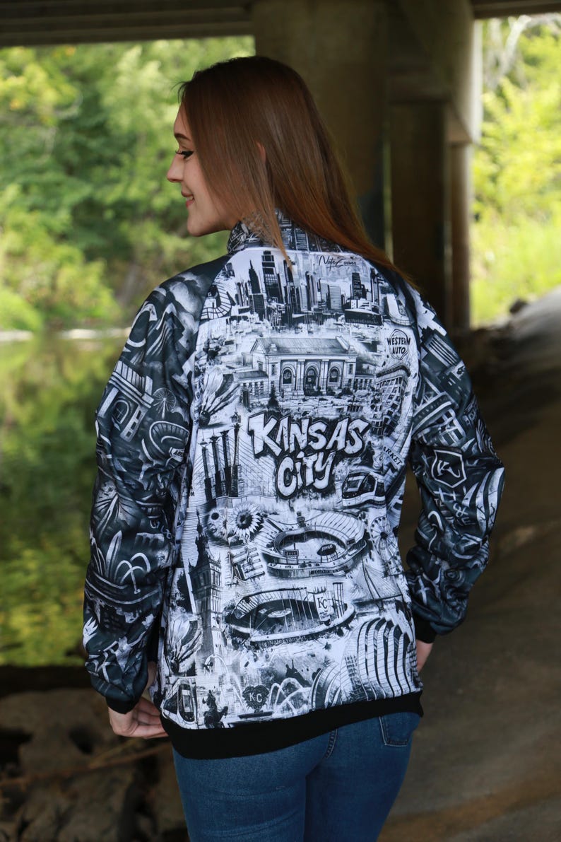 May include: A black and white zip-up jacket featuring a detailed graphic of Kansas City landmarks and the city's name. The jacket has a black collar and cuffs, and is worn with blue jeans.