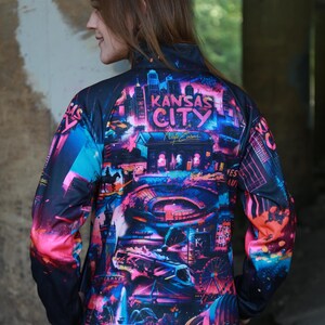 May include: A black zip-up jacket featuring a vibrant, colorful graphic of the Kansas City skyline. The design includes the words "Kansas City" and other city landmarks in shades of pink, blue, and purple.