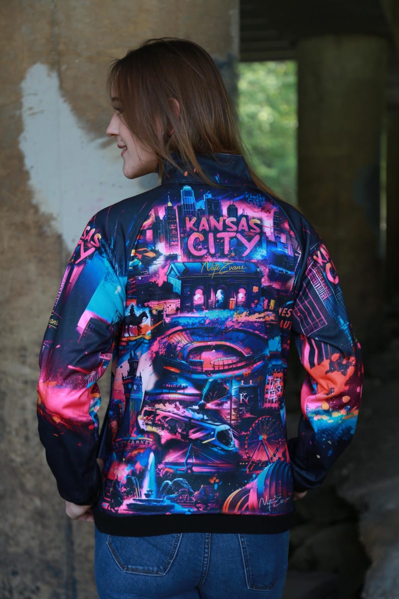 May include: A black zip-up jacket featuring a vibrant, colorful graphic of the Kansas City skyline. The design includes the words "Kansas City" and other city landmarks in shades of pink, blue, and purple.