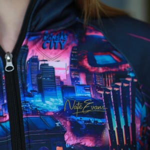 May include: A zip-up hoodie featuring a vibrant cityscape design in shades of blue, pink, and purple. The words "Kansas City" are visible at the top, with "Nate Evans" in gold script below. The hoodie has a black zipper.
