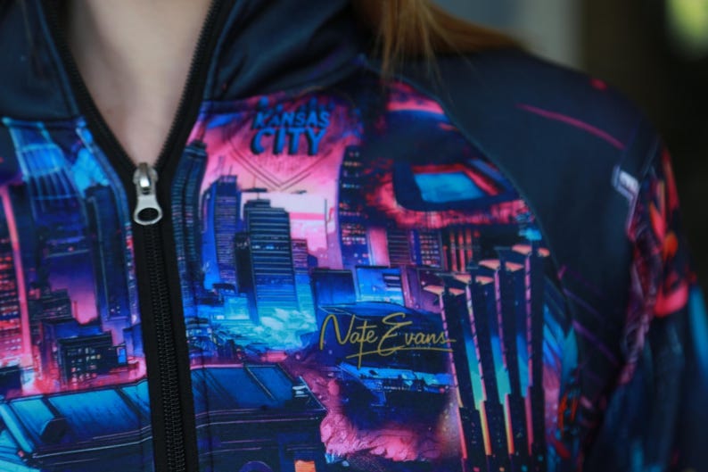 May include: A zip-up hoodie featuring a vibrant cityscape design in shades of blue, pink, and purple. The words "Kansas City" are visible at the top, with "Nate Evans" in gold script below. The hoodie has a black zipper.