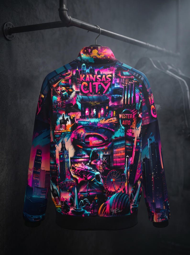 May include: A vibrant bomber jacket featuring a colorful graphic of Kansas City landmarks. The design includes the city's skyline, Ferris wheel, and the words "Kansas City" in bright pink. The jacket is hanging on a metal rack.