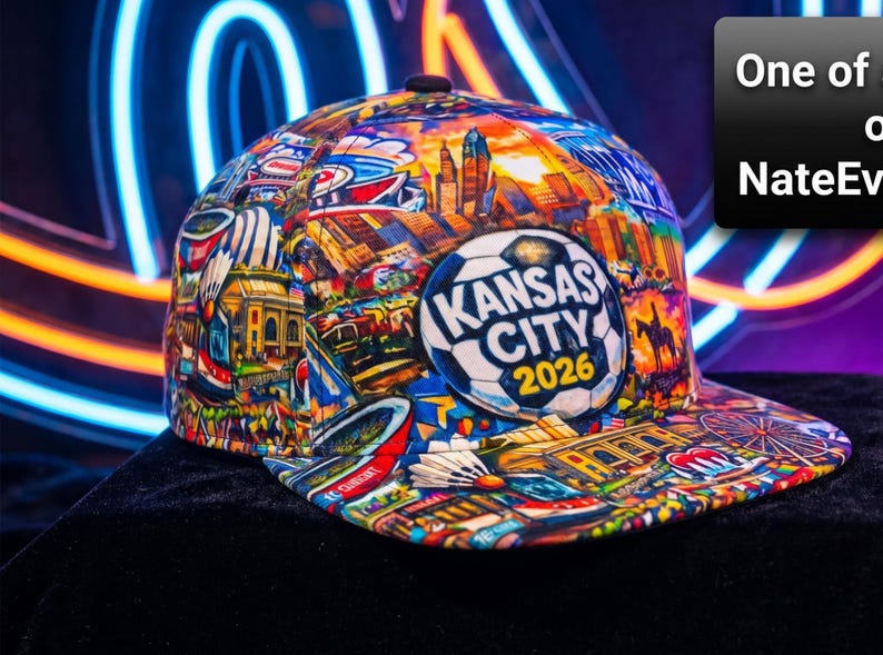 May include: A colorful baseball cap featuring a collage of Kansas City landmarks and the text "Kansas City 2026" on a soccer ball emblem. The cap's design includes buildings, a Ferris wheel, and other city elements.