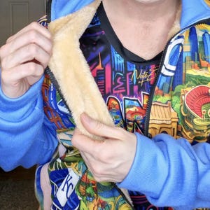 May include: A blue zip-up jacket with a colorful cityscape design. The jacket has a light tan, fuzzy lining and blue ribbed cuffs. The inner shirt has a graphic design with the name "Nate" on it.