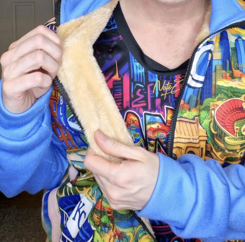 May include: A blue zip-up jacket with a colorful cityscape design. The jacket has a light tan, fuzzy lining and blue ribbed cuffs. The inner shirt has a graphic design with the name "Nate" on it.