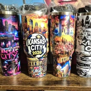 May include: Four stainless steel travel mugs with black lids. Each mug features a colorful or black and white design of the Kansas City skyline. One mug has a soccer ball graphic with "Kansas City 2026" text. The mugs are on a wooden surface.