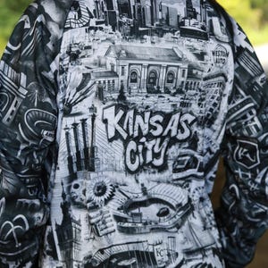 May include: Black and white jacket featuring a detailed graphic of Kansas City landmarks and the city's name in bold letters. The design includes iconic buildings, bridges, and architectural elements, creating a unique urban-themed garment.