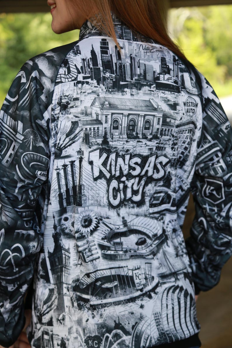 May include: Black and white jacket featuring a detailed graphic of Kansas City landmarks and the city's name in bold letters. The design includes iconic buildings, bridges, and architectural elements, creating a unique urban-themed garment.