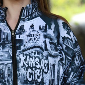May include: A black and white zip-up jacket featuring a cityscape design with the words "Kansas City" in large letters. The jacket includes the "Western Auto" logo and the name "Nate Evans".