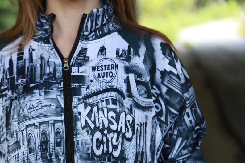 May include: A black and white zip-up jacket featuring a cityscape design with the words "Kansas City" in large letters. The jacket includes the "Western Auto" logo and the name "Nate Evans".