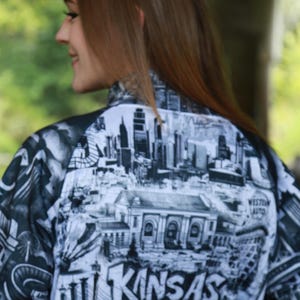 May include: A black and white jacket featuring a detailed graphic of the Kansas City skyline and the city's name in bold lettering. The jacket's design includes iconic landmarks and architectural elements.