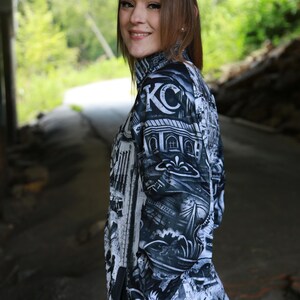 May include: Black and white zip-up jacket with a detailed graphic print featuring architectural elements and the letters "KC". The jacket has a high collar and black trim. The background is a blurred outdoor setting.