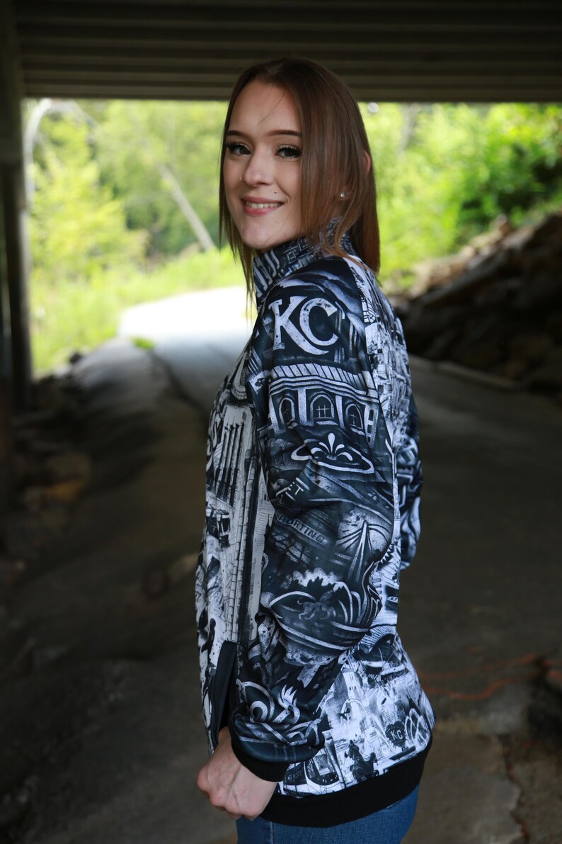 May include: Black and white zip-up jacket with a detailed graphic print featuring architectural elements and the letters "KC". The jacket has a high collar and black trim. The background is a blurred outdoor setting.