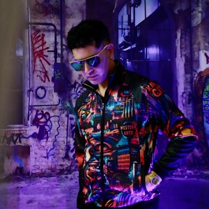 May include: A black zip-up jacket with a colorful cityscape design, including the text "Western Auto" and "Kansas City". The jacket has a black collar and sleeves. The person is wearing gold-rimmed sunglasses and a gold watch.