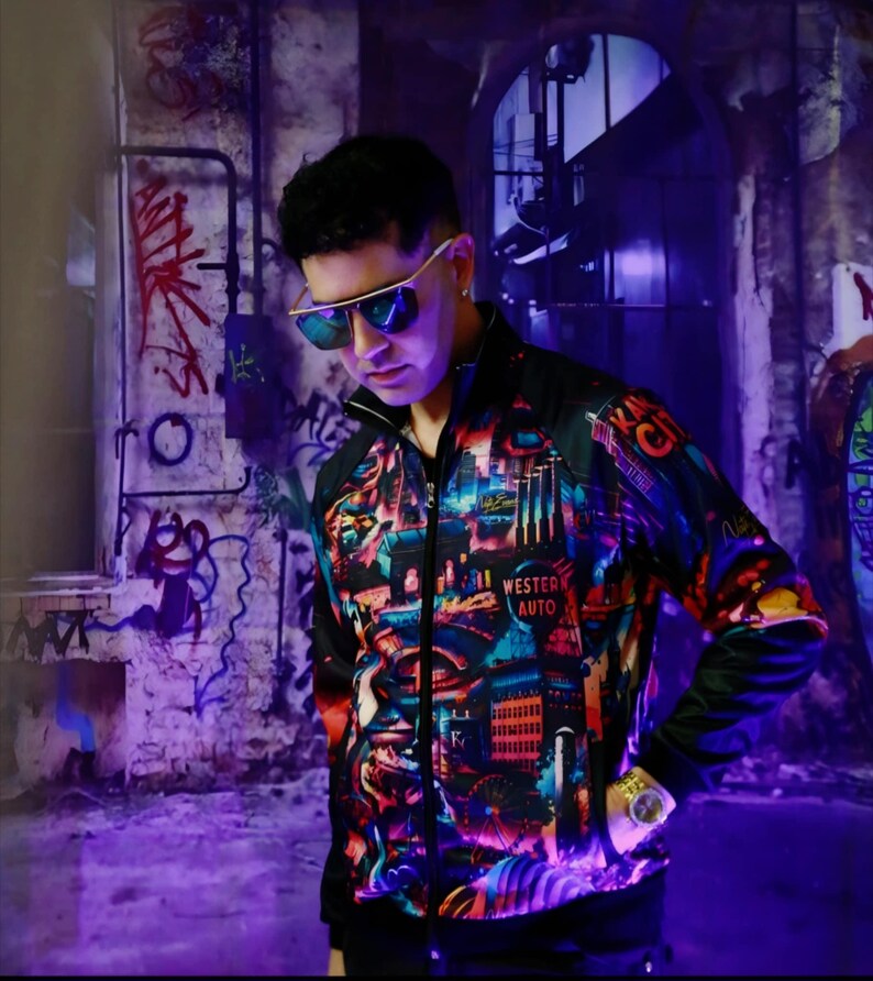 May include: A black zip-up jacket with a colorful cityscape design, including the text "Western Auto" and "Kansas City". The jacket has a black collar and sleeves. The person is wearing gold-rimmed sunglasses and a gold watch.