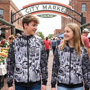 May include: Two people wearing matching black and white zip-up jackets with a cityscape design and the text "Kansas City". They are walking in front of a "City Market" archway, surrounded by produce stands.
