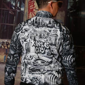 May include: A black and white zip-up jacket featuring a detailed graphic of Kansas City landmarks and the city's name. The jacket has a black collar and cuffs, and the design includes buildings, a Ferris wheel, and other city elements.