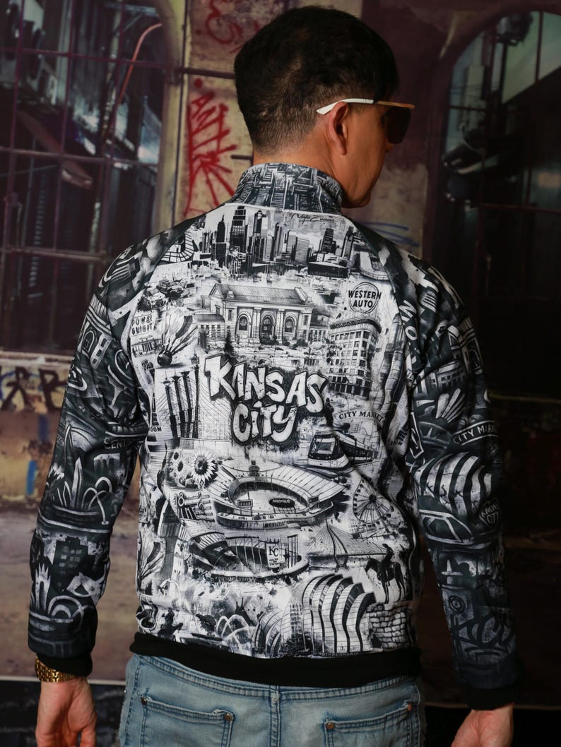 May include: A black and white zip-up jacket featuring a detailed graphic of Kansas City landmarks and the city's name. The jacket has a black collar and cuffs, and the design includes buildings, a Ferris wheel, and other city elements.