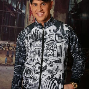 May include: A black and white zip-up jacket featuring a detailed graphic of Kansas City landmarks, including the words "Kansas City" and "Western Auto". The jacket has a black collar, cuffs, and hem. The person is wearing a gold watch.