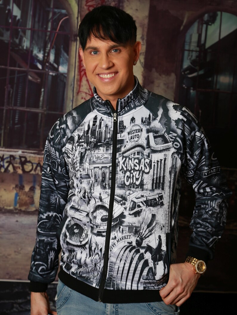 May include: A black and white zip-up jacket featuring a detailed graphic of Kansas City landmarks, including the words "Kansas City" and "Western Auto". The jacket has a black collar, cuffs, and hem. The person is wearing a gold watch.