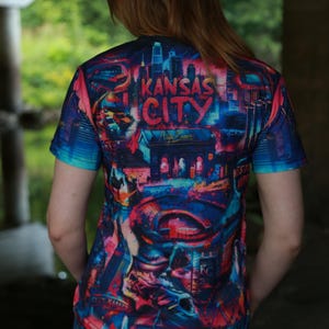 K&ouml;nnte beinhalten: Ein kurz&auml;rmeliges T-Shirt mit einem lebendigen, farbenfrohen Grafikdesign mit den Worten "Kansas City" in einer stilisierten Schriftart. Das Design enth&auml;lt Stadtlandschaften in Blau-, Rot- und Lilat&ouml;nen. Das T-Shirt wird von einer Person getragen.