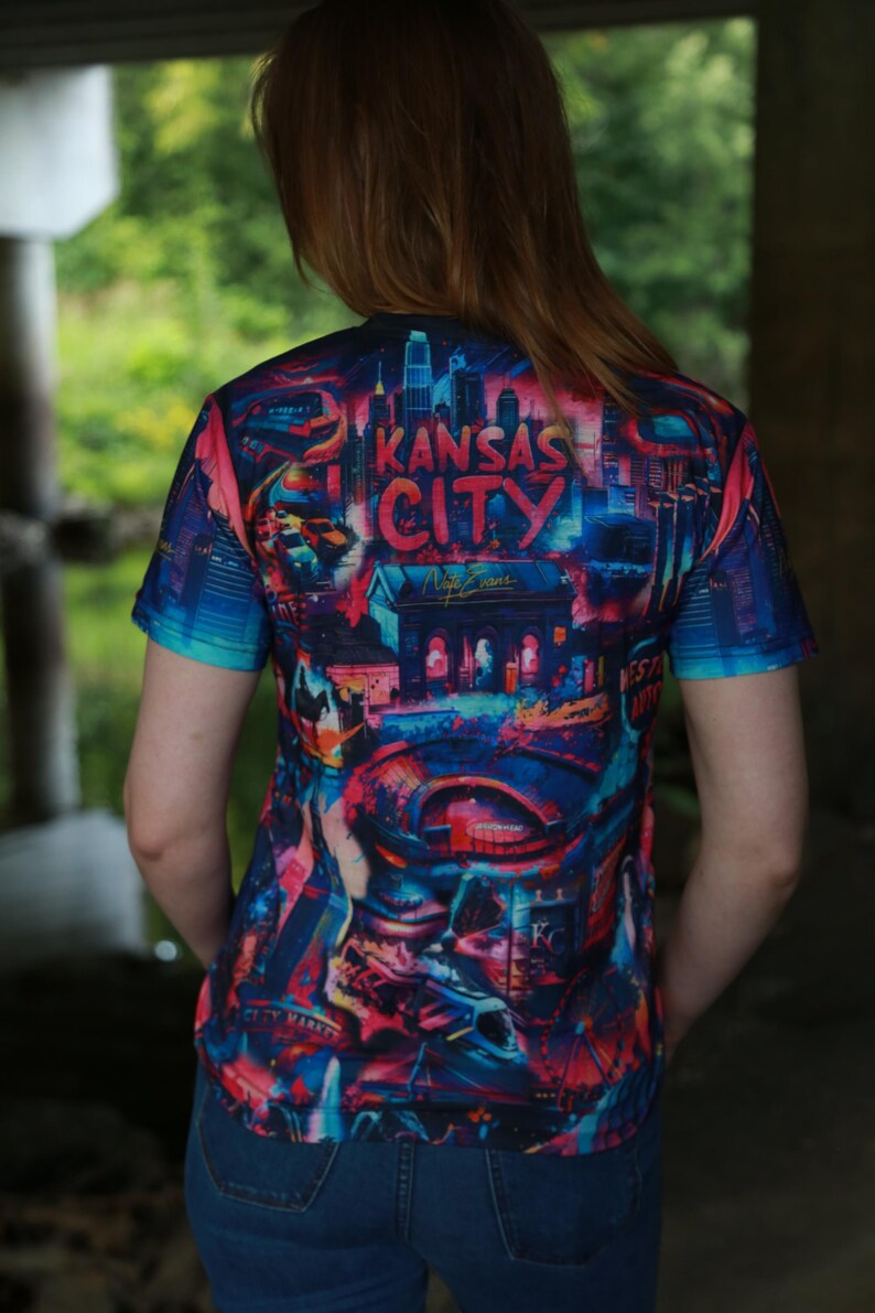 K&ouml;nnte beinhalten: Ein kurz&auml;rmeliges T-Shirt mit einem lebendigen, farbenfrohen Grafikdesign mit den Worten "Kansas City" in einer stilisierten Schriftart. Das Design enth&auml;lt Stadtlandschaften in Blau-, Rot- und Lilat&ouml;nen. Das T-Shirt wird von einer Person getragen.