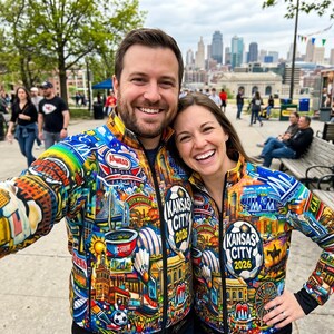 Puede incluir: Una pareja sonr&iacute;e, vistiendo chaquetas con cremallera a juego con ilustraciones coloridas de los lugares emblem&aacute;ticos de Kansas City y el texto "Kansas City 2026". Las chaquetas presentan un dise&ntilde;o de arco&iacute;ris e iconos de la ciudad.