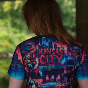 K&ouml;nnte beinhalten: Ein kurz&auml;rmeliges T-Shirt mit einer farbenfrohen Grafik der Skyline von Kansas City. Das Design zeigt den Namen der Stadt in fetten Buchstaben, mit einer Mischung aus roten, blauen und rosa Farbt&ouml;nen. Das T-Shirt wird von einer Person mit langen, rotbraunen Haaren getragen.