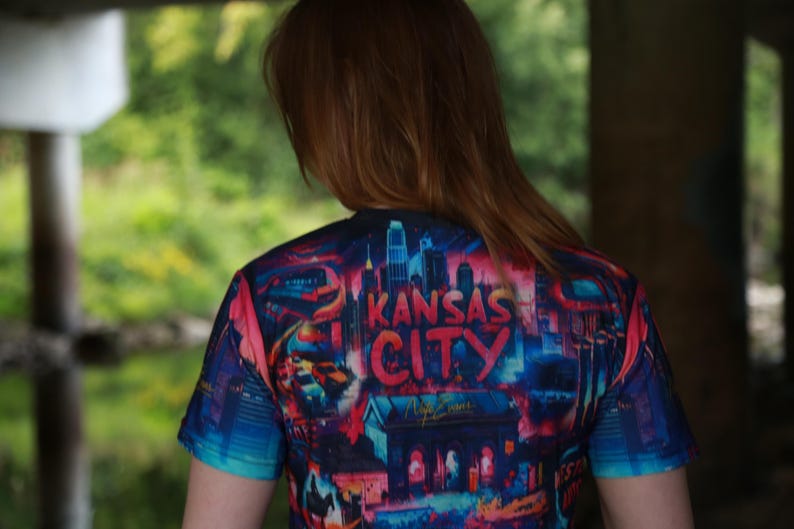 K&ouml;nnte beinhalten: Ein kurz&auml;rmeliges T-Shirt mit einer farbenfrohen Grafik der Skyline von Kansas City. Das Design zeigt den Namen der Stadt in fetten Buchstaben, mit einer Mischung aus roten, blauen und rosa Farbt&ouml;nen. Das T-Shirt wird von einer Person mit langen, rotbraunen Haaren getragen.