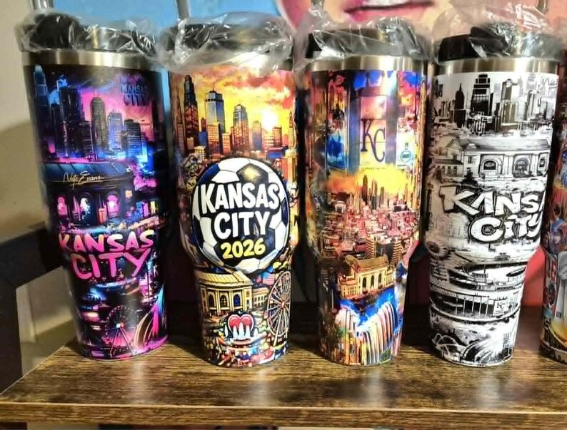 Może przedstawiać: Cztery kubki podr&oacute;żne ze stali nierdzewnej z czarnymi pokrywkami. Każdy kubek ma kolorowy lub czarno-biały wz&oacute;r panoramy Kansas City. Jeden kubek ma grafikę piłki nożnej z tekstem "Kansas City 2026". Kubki stoją na drewnianej powierzchni.