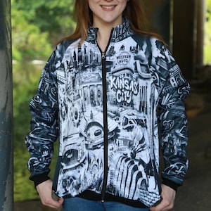 May include: A zip-up jacket featuring a black and white cityscape design with the words "Kansas City" prominently displayed. The jacket has a black collar, cuffs, and zipper, and is worn with blue jeans.
