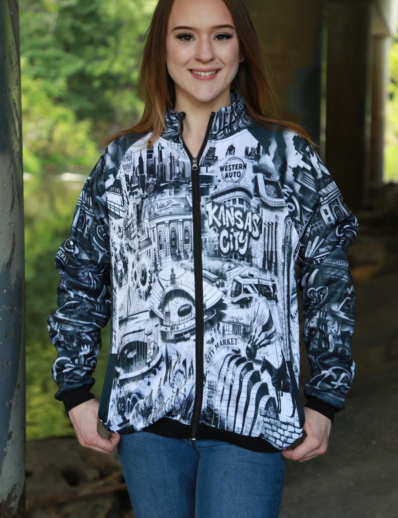 May include: A zip-up jacket featuring a black and white cityscape design with the words "Kansas City" prominently displayed. The jacket has a black collar, cuffs, and zipper, and is worn with blue jeans.