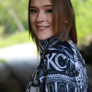 May include: A black and white patterned mock neck shirt with the letters "KC" prominently displayed. The shirt features a cityscape design and is worn by a person with brown hair.