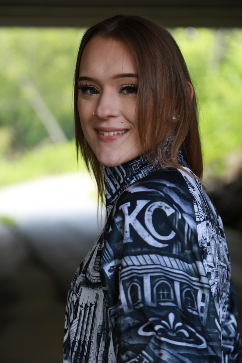 May include: A black and white patterned mock neck shirt with the letters "KC" prominently displayed. The shirt features a cityscape design and is worn by a person with brown hair.